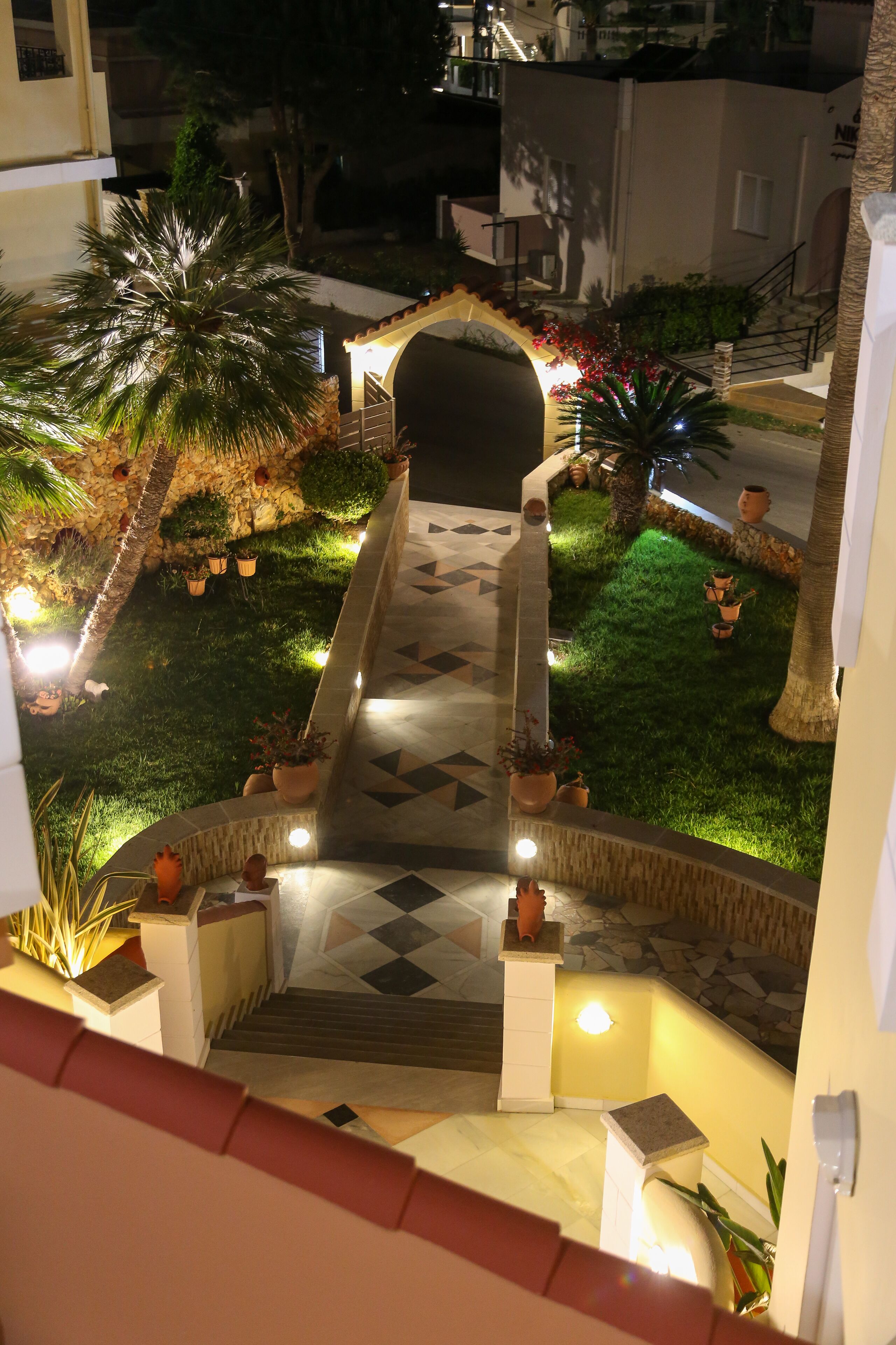 A well-lit outdoor area with a stone pathway leading to a covered seating area, surrounded by potted plants and palm trees.