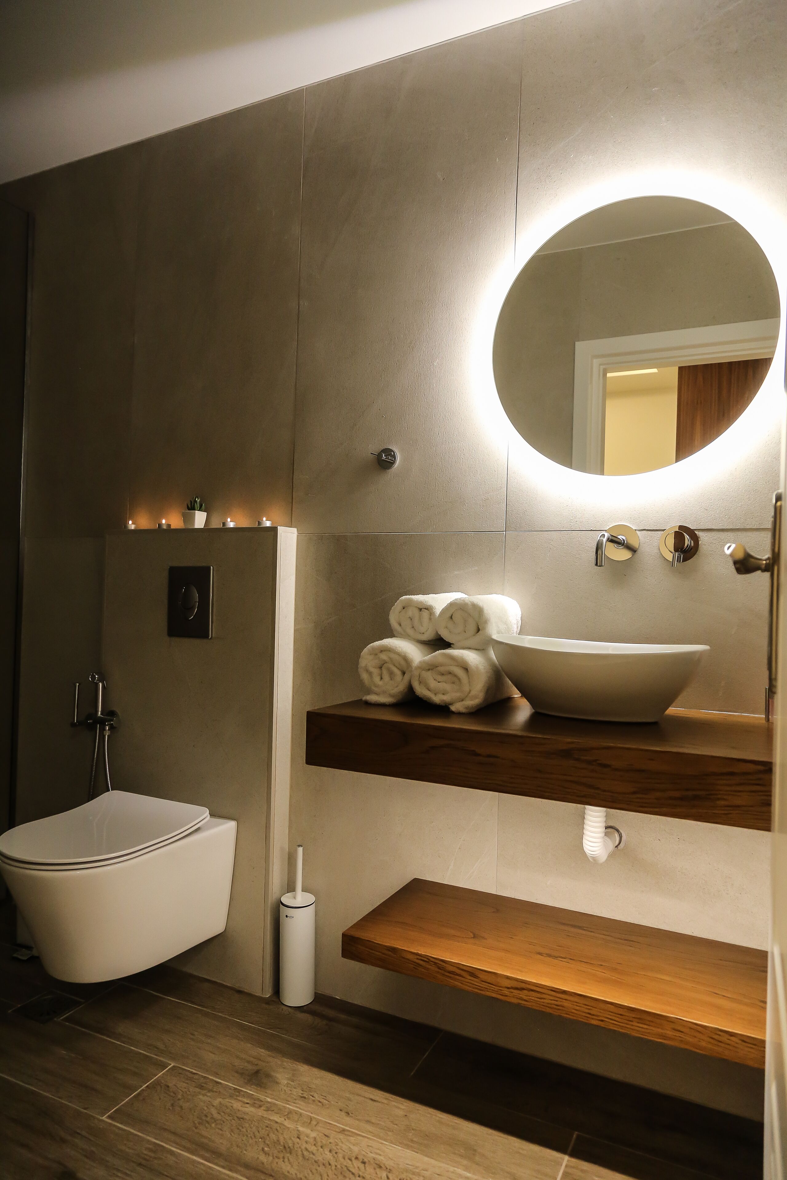 Un baño moderno con inodoro integrado, un espejo redondo con luz, un estante de madera con toallas y un lavabo blanco.