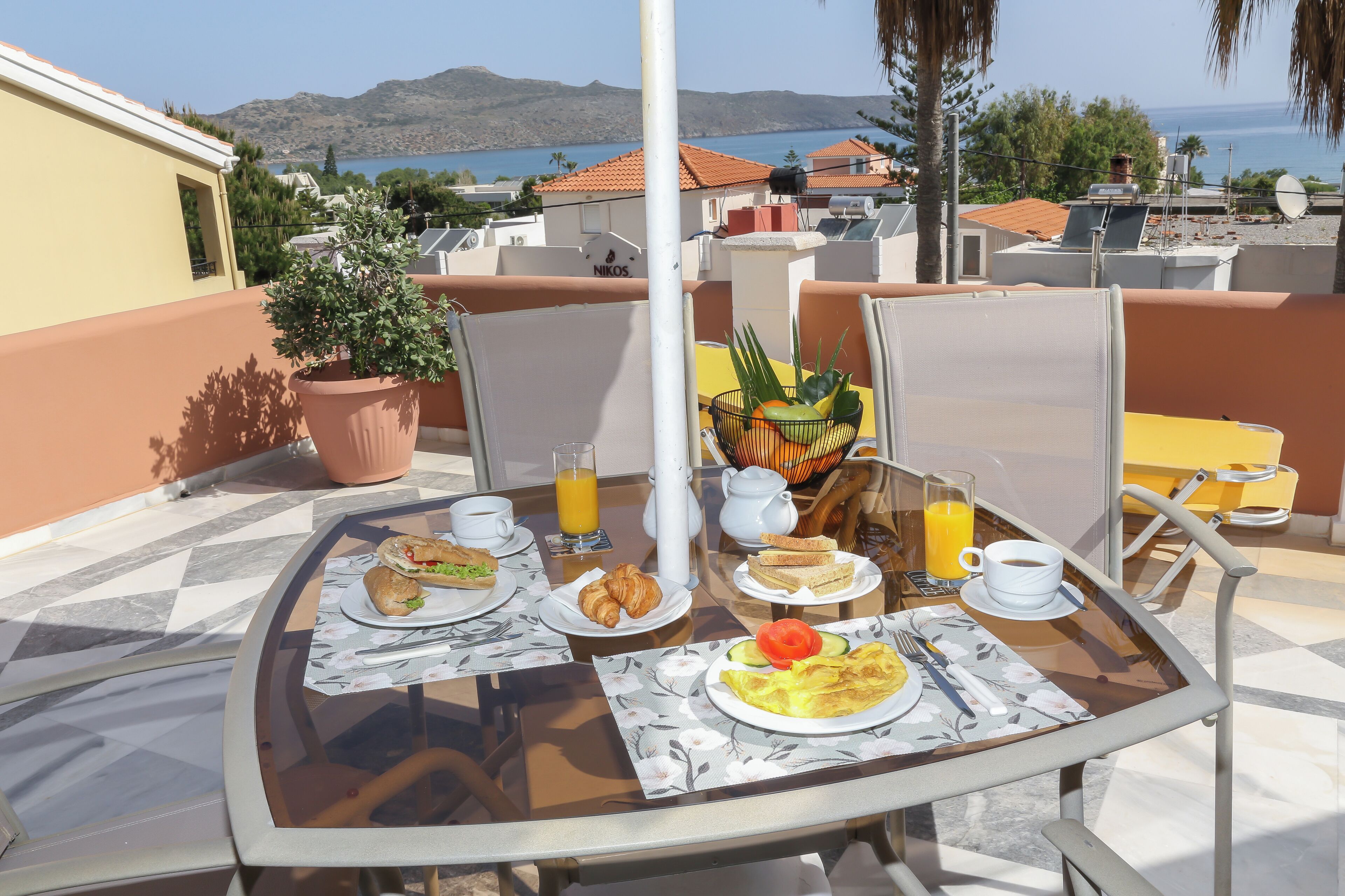A breakfast table with croissants, a plate of eggs, and a glass of orange juice on a terrace with a view of the sea and mountains.