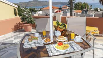 A breakfast table with croissants, a plate of eggs, and a glass of orange juice on a terrace with a view of the sea and mountains.