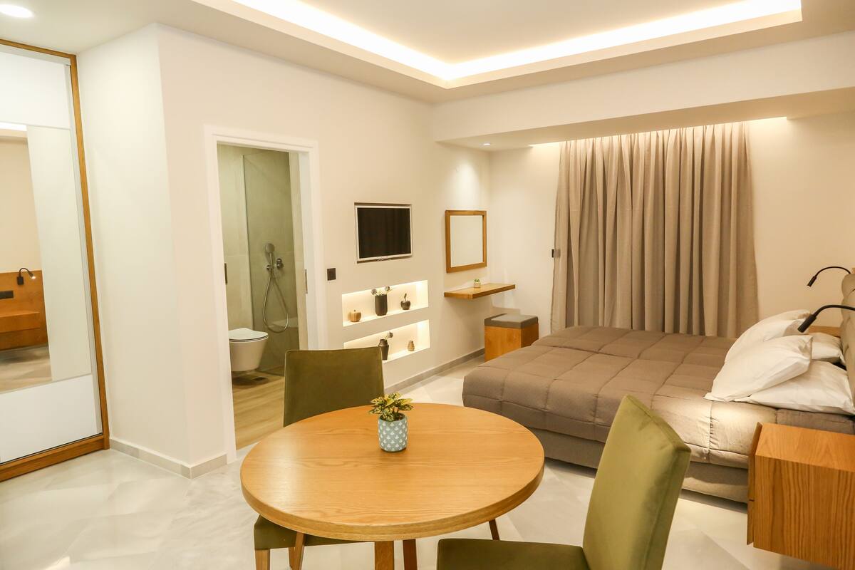 Ein modernes Hotelzimmer mit einem Bett, einem runden Holztisch mit Stühlen, einer kleinen Topfpflanze, einem an der Wand montierten Fernseher und einem Fenster mit Vorhängen.