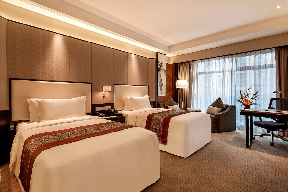 Habitació d'hotel amb dos llits, una taula de treball, una cadira, una làmpada i una finestra gran amb cortines.
