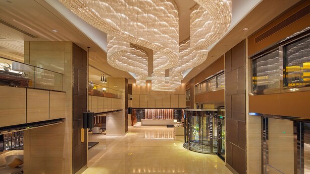 En rummelig lobby med en moderne lysekrone, glas elevator og en receptionsdisk.