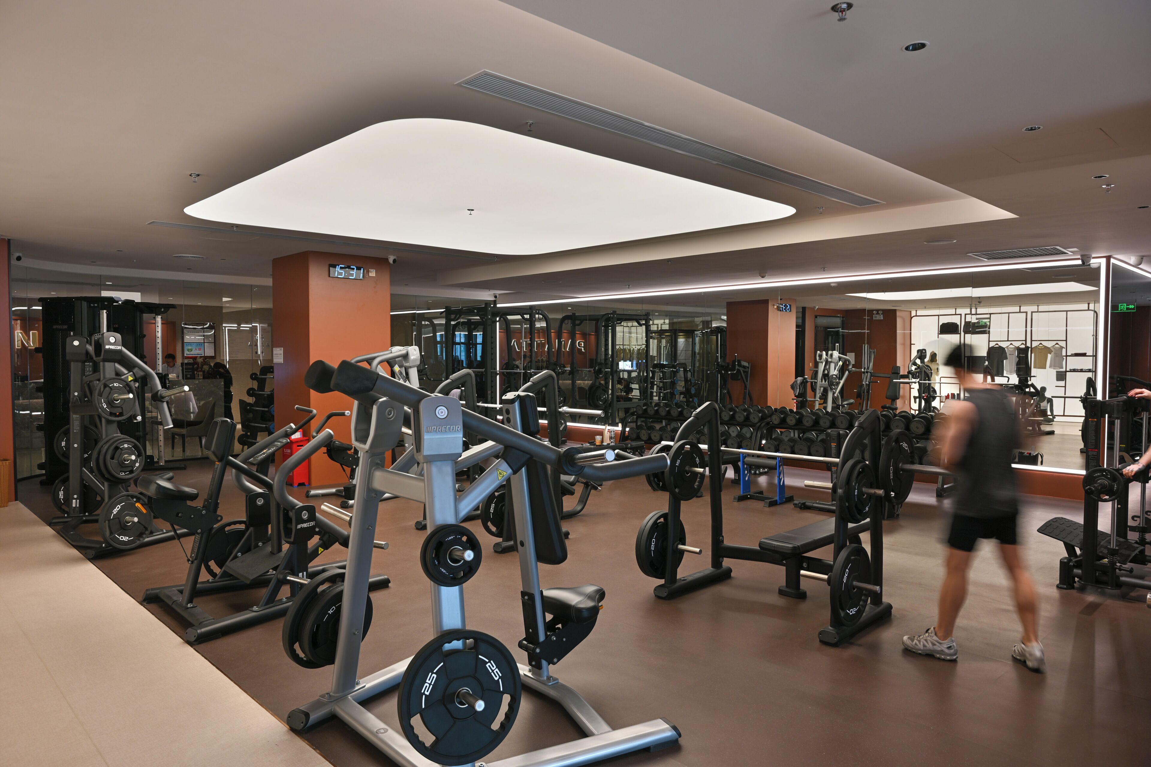 A modern gym with various weightlifting equipment, a digital clock, and a large mirror.