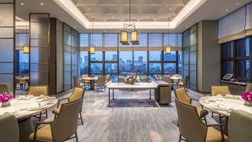 A spacious dining area with high ceilings, large windows offering a city view, and modern lighting fixtures.