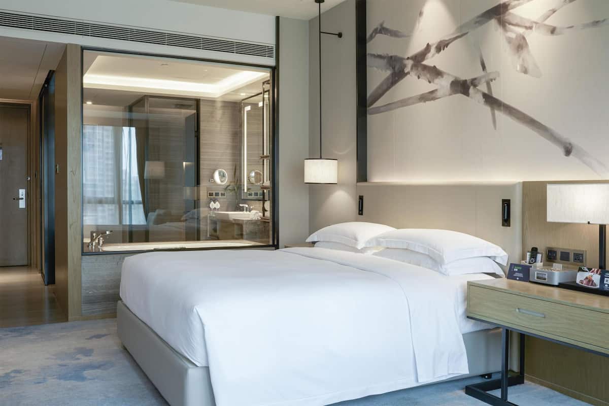 A modern hotel room with a large bed, a glass shower area, and a wall-mounted lamp.