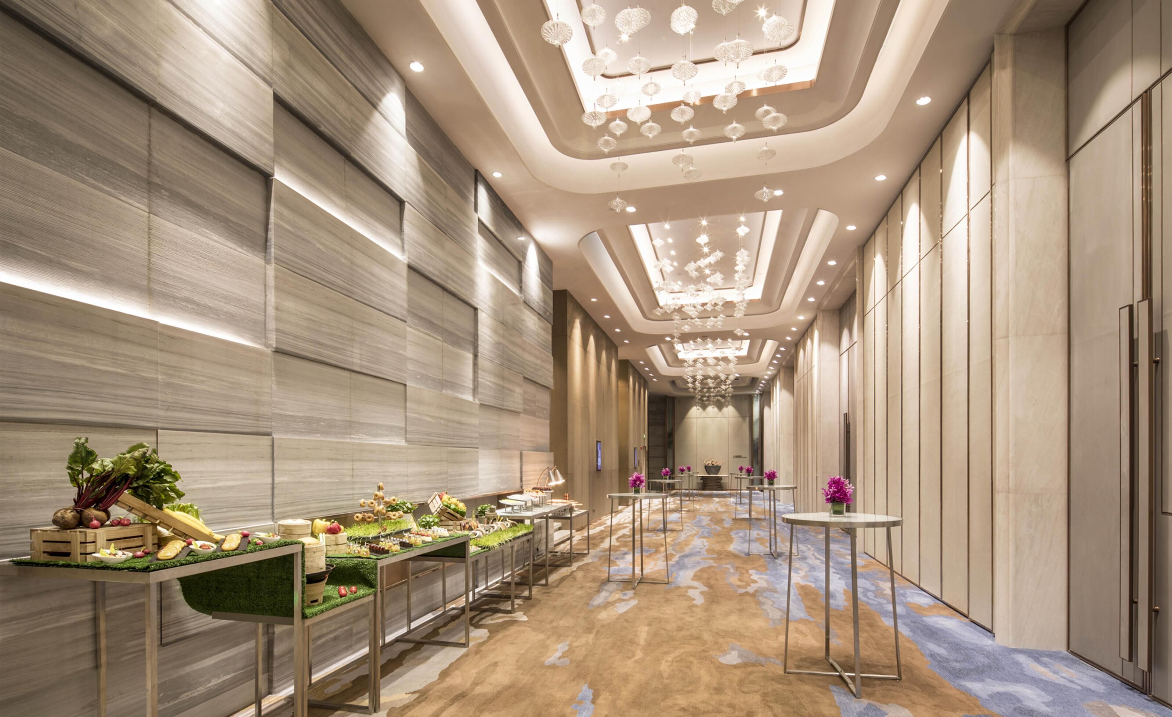 Un largo y estrecho pasillo de hotel con un pasarela central, luces decorativas en el techo y un área de bufé con frutas frescas a la izquierda.