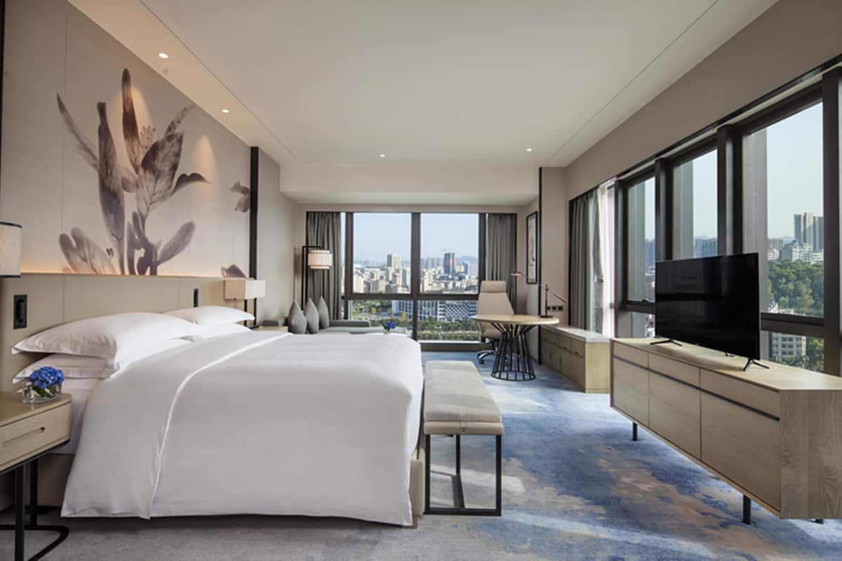 A modern hotel room with a large bed, a desk, a chair, a TV, and a city view through large windows.