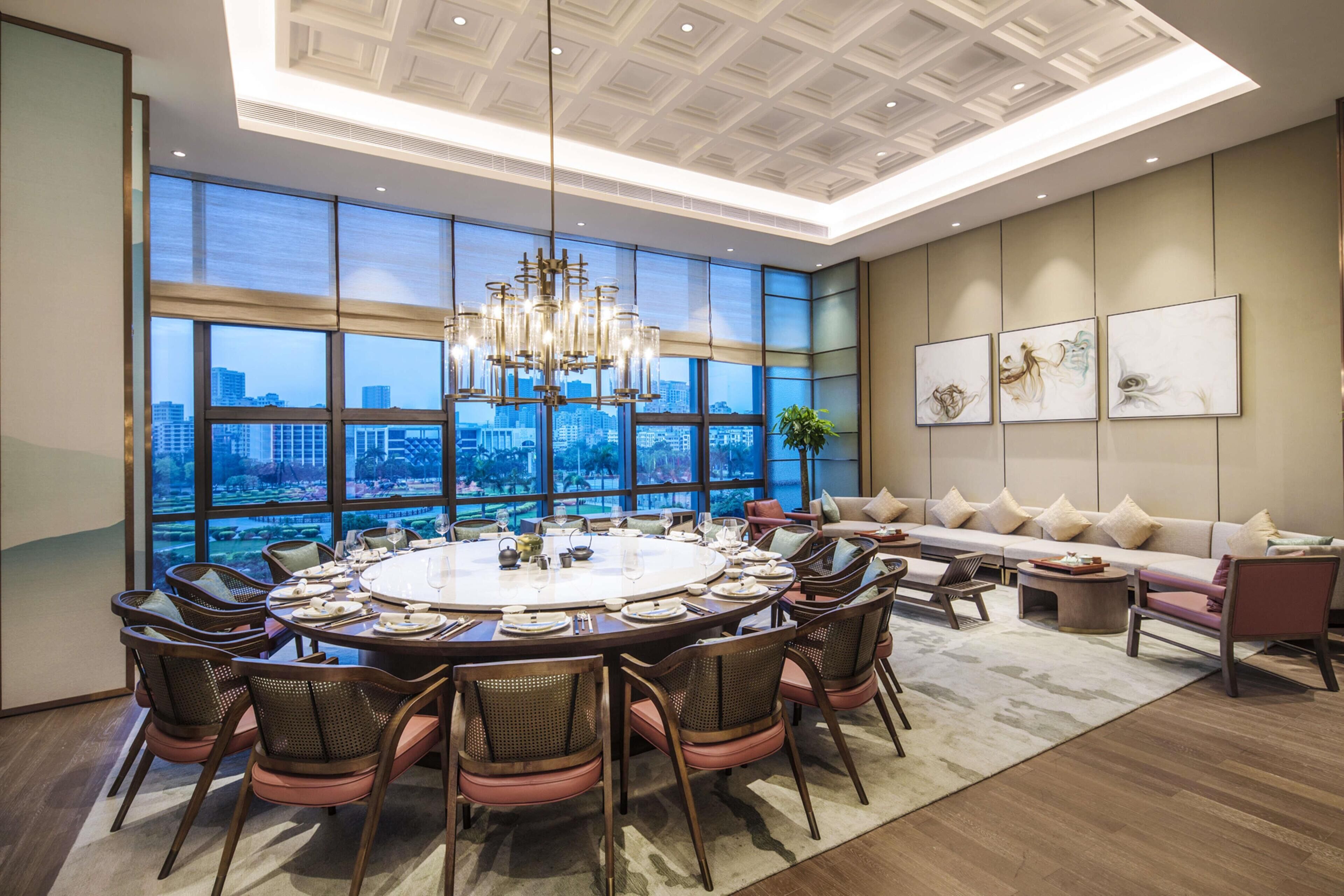 A spacious room with a large round table, comfortable seating, and a city view through large windows.