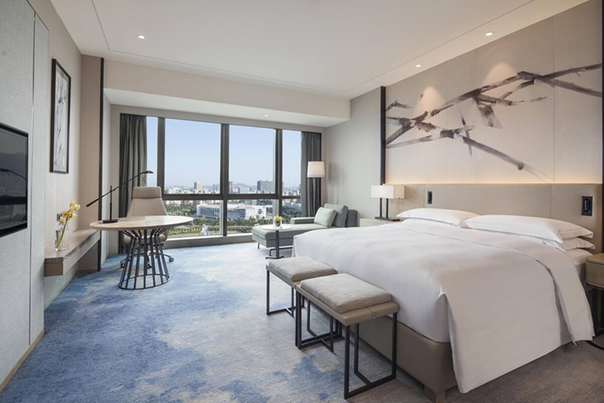 A modern hotel room with a large bed, a desk, a chair, and a view of the city.