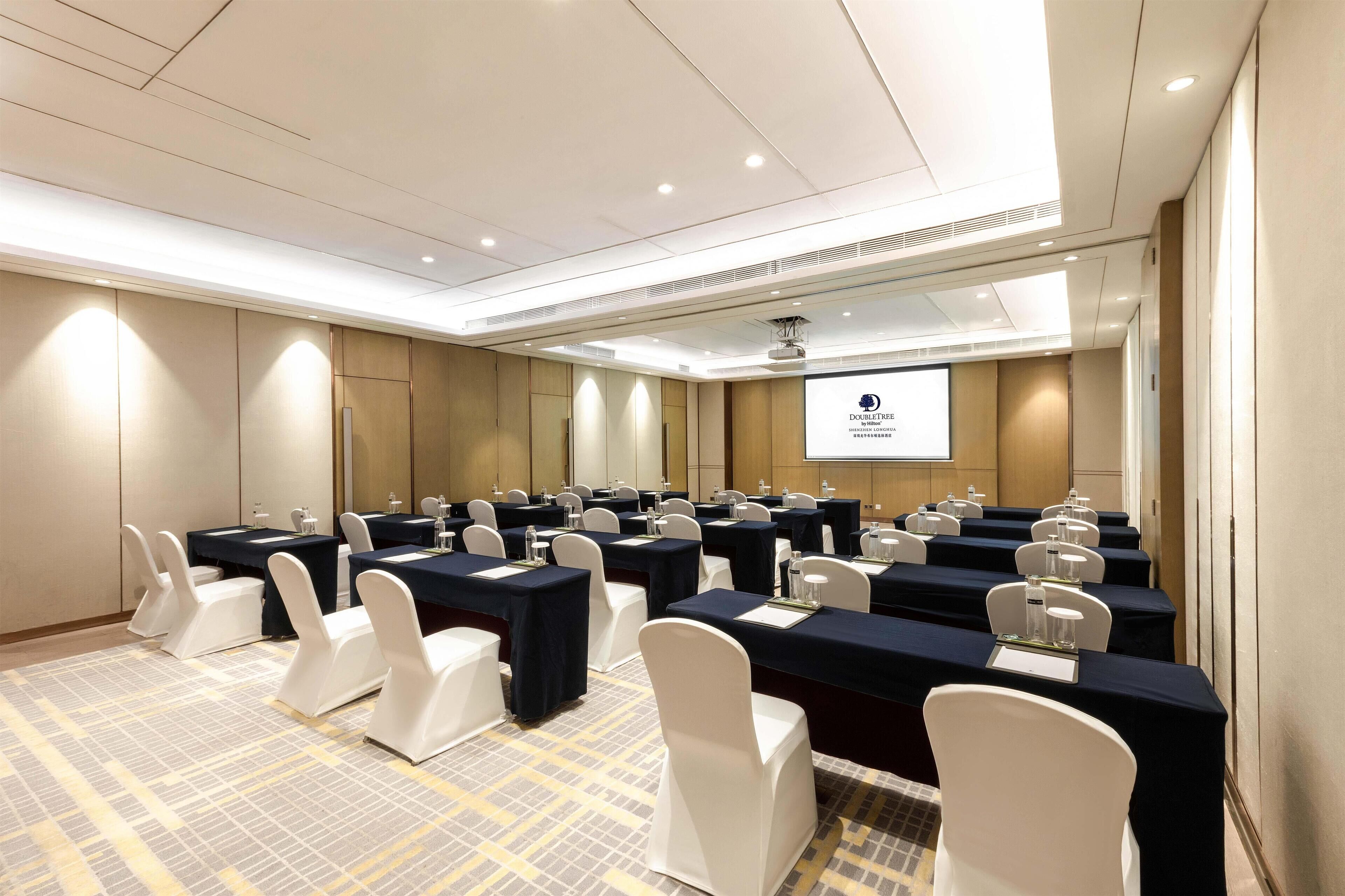 A conference room with multiple tables and chairs, a projector screen, and a carpeted floor.