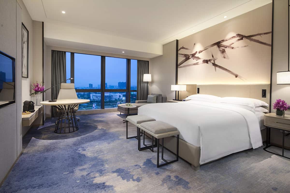 A modern hotel room with a large bed, a desk, and a view of the cityscape.