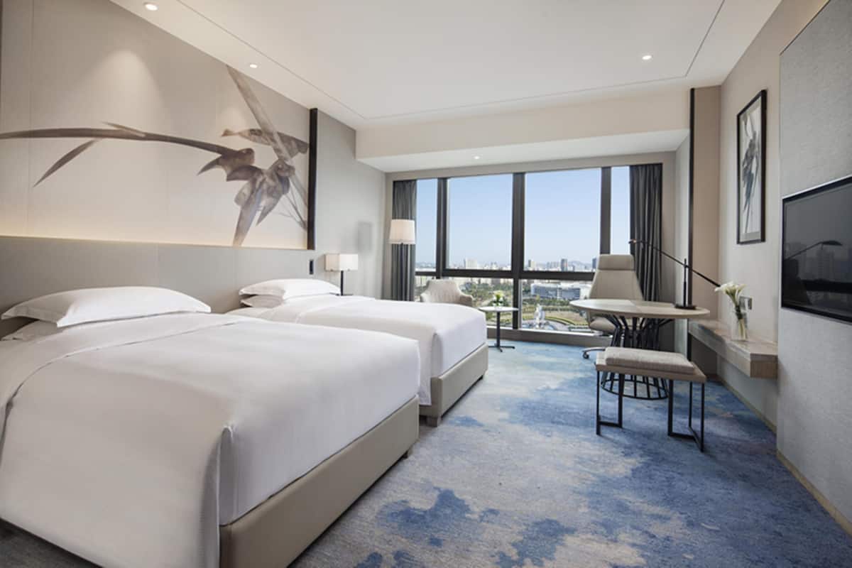 A hotel room with two beds, a small table, a chair, a TV, and a large window offering a city view.