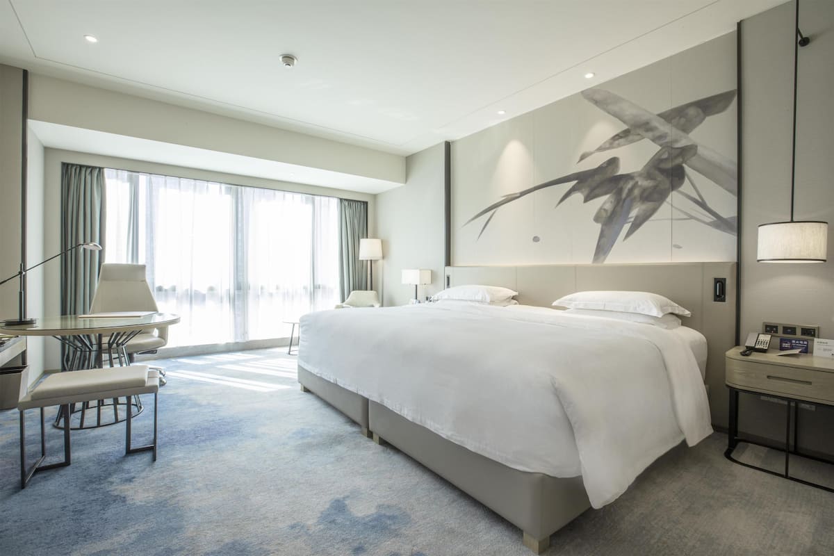 A modern hotel room with a large bed, a desk with a chair, a bench, and a large window with curtains.