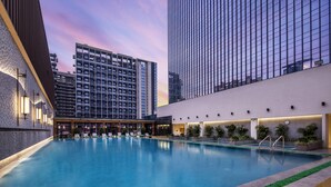 Zona de piscina moderna de hotel con vistas a edificios altos, piscina bien iluminada y sillones de descanso.