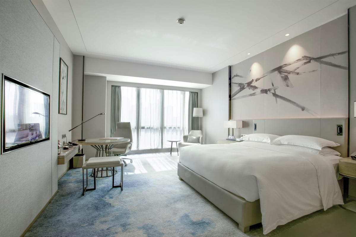 A modern hotel room with a large bed, a desk with a chair, a small table, and a large window with curtains.