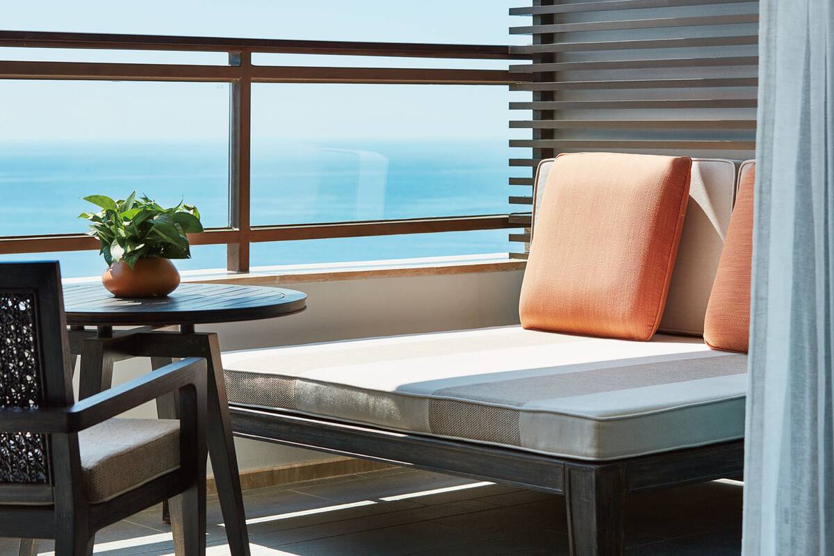 A balcony with a cushioned seating area, a small table with a potted plant, and a view of the ocean.
