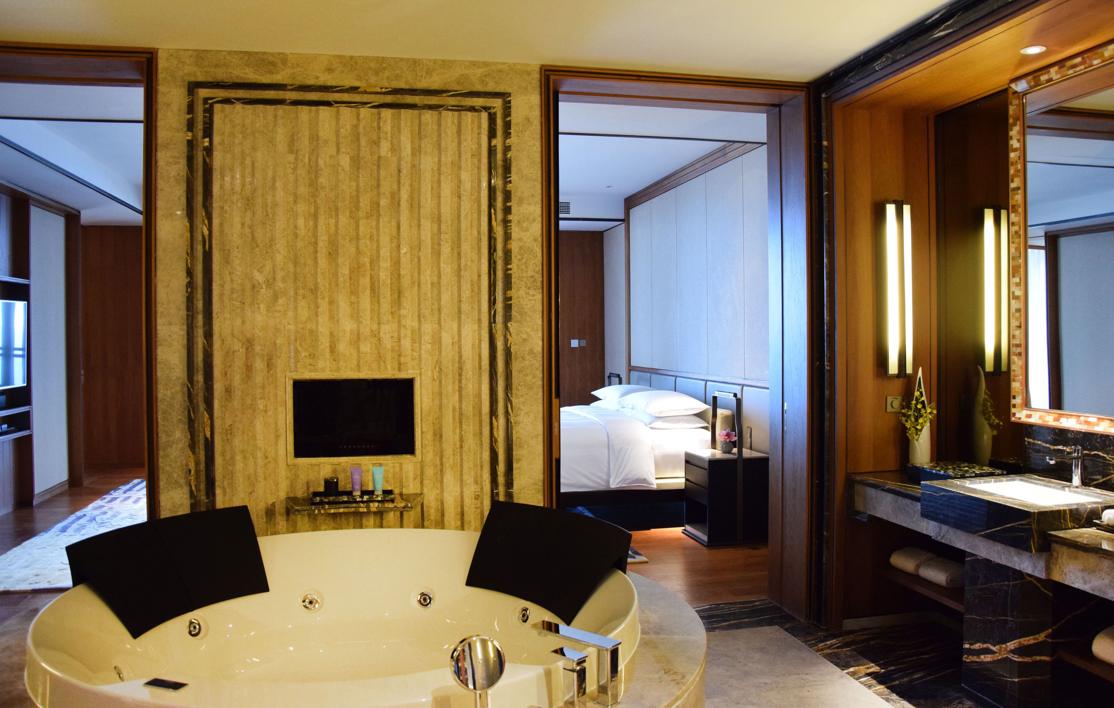 A modern hotel room with a large bathtub, a bed, a television, and a bathroom with a sink and mirror.