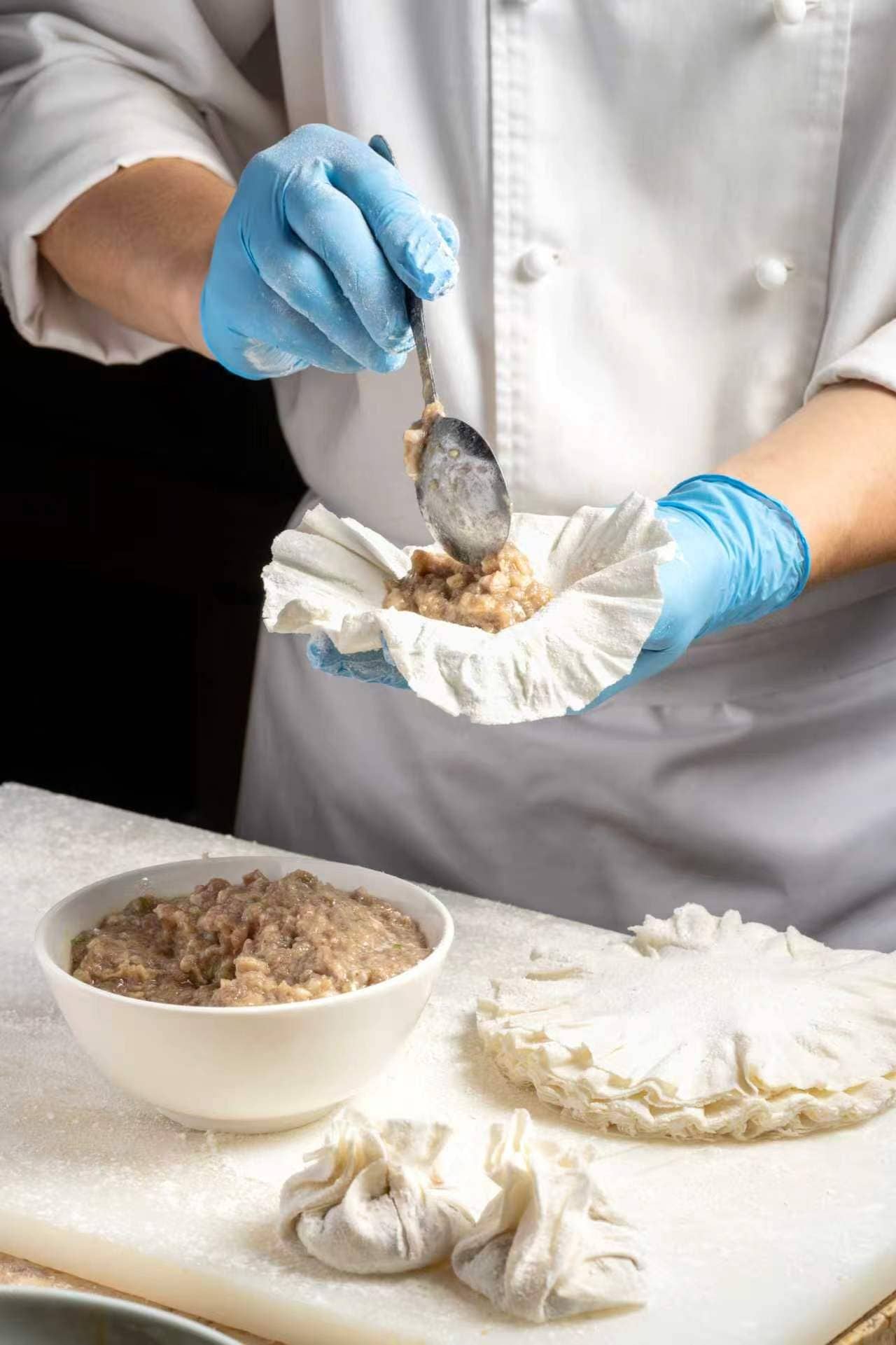 A person in a white chef's coat and blue gloves preparing food, with a bowl of mixture and a piece of dough on the counter.