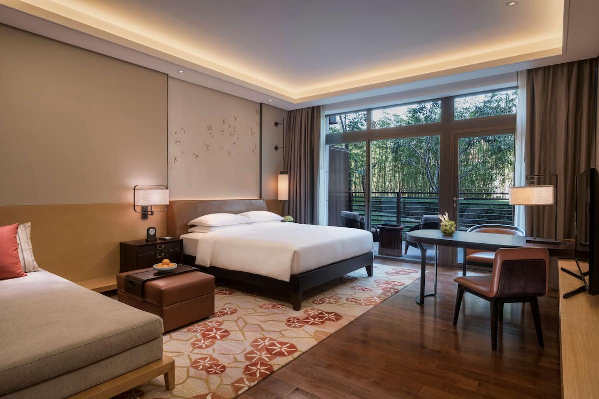 Ein Hotelzimmer mit einem großen Bett, einem Schreibtisch, einem Sessel, einem kleinen Tisch und einem Balkon mit Blick auf Bäume.