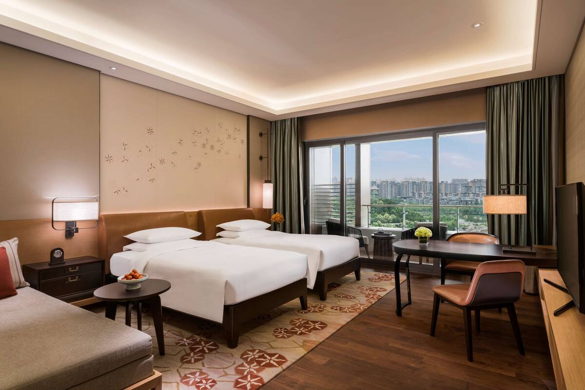 Ein Hotelzimmer mit zwei Betten, einem Schreibtisch, einem Stuhl, einem Fernseher und Blick auf die Stadt.