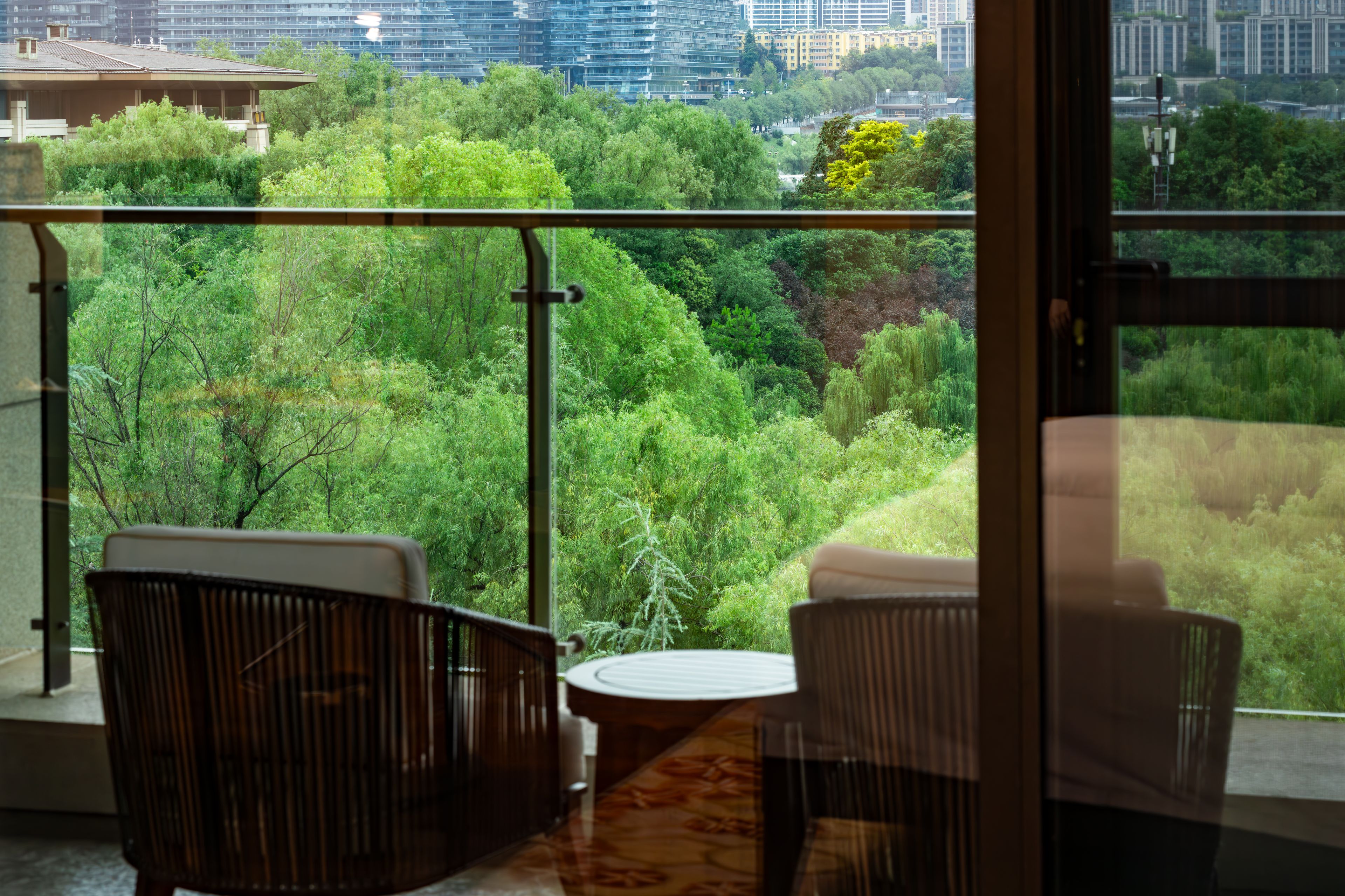 Un balcón con dos sillas y una mesita, con vistas a un paisaje verde exuberante y edificios urbanos.