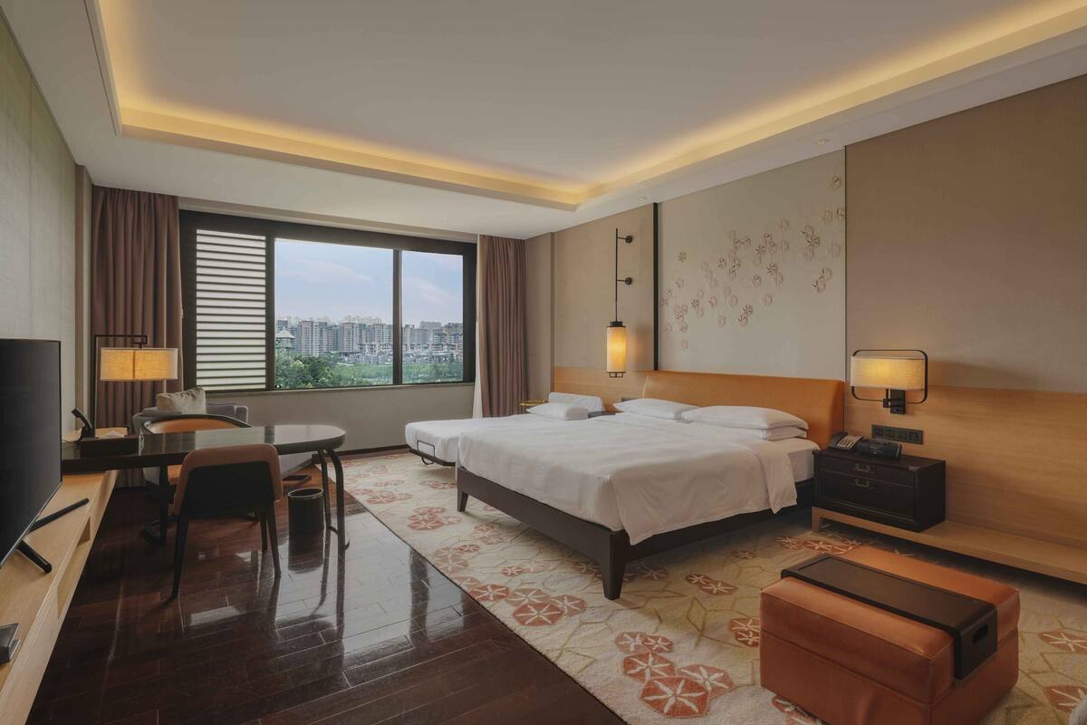 Ein Hotelzimmer mit einem großen Bett, einem Schreibtisch mit Stuhl, einem Fernseher und Blick auf die Stadt durch das Fenster.