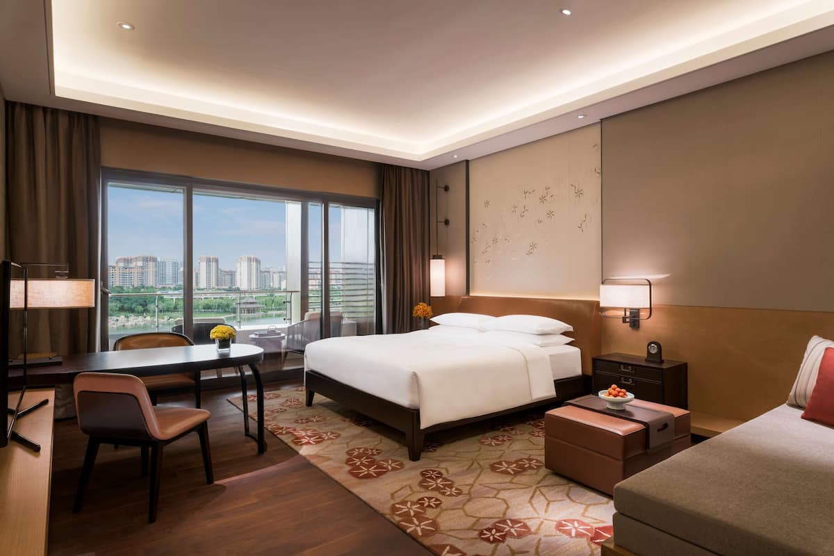 Ein Hotelzimmer mit einem großen Bett, einem Schreibtisch mit Stuhl, einem kleinen Tisch mit Vase, einem Nachttisch mit Lampe und Blick auf die Stadt durch große Fenster.