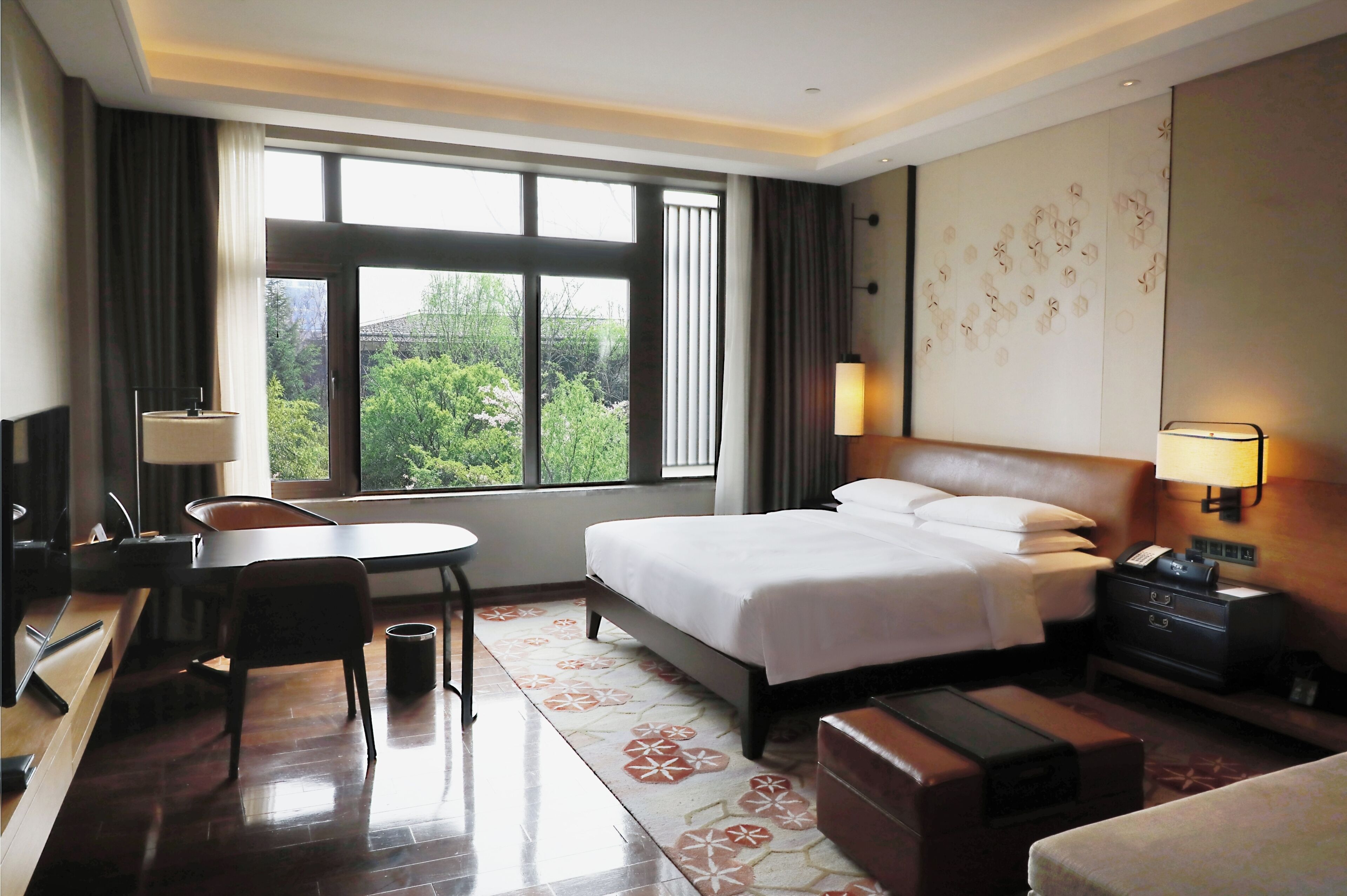 Habitación de hotel con cama, escritorio, silla, lámpara y vistas al verdor exterior.