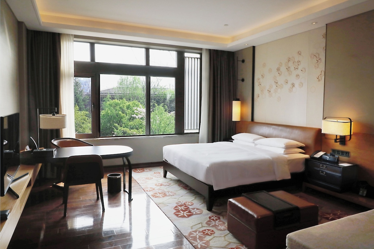 A hotel room with a bed, desk, chair, lamp, and a view of greenery outside.