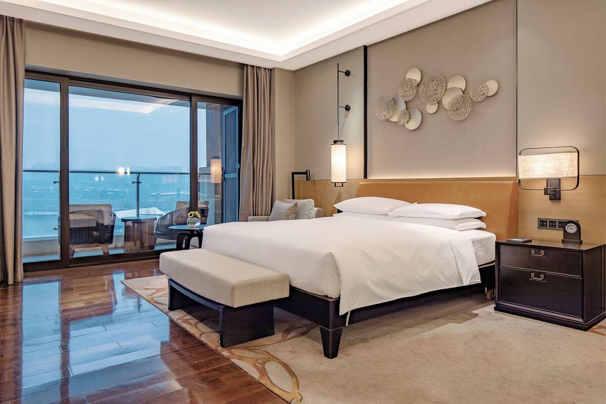 Ein modernes Hotelzimmer mit einem großen Bett, einem Nachttisch, einer Bank und einem Blick auf die Stadt durch raumhohe Fenster, sowie einem Wanddekor.