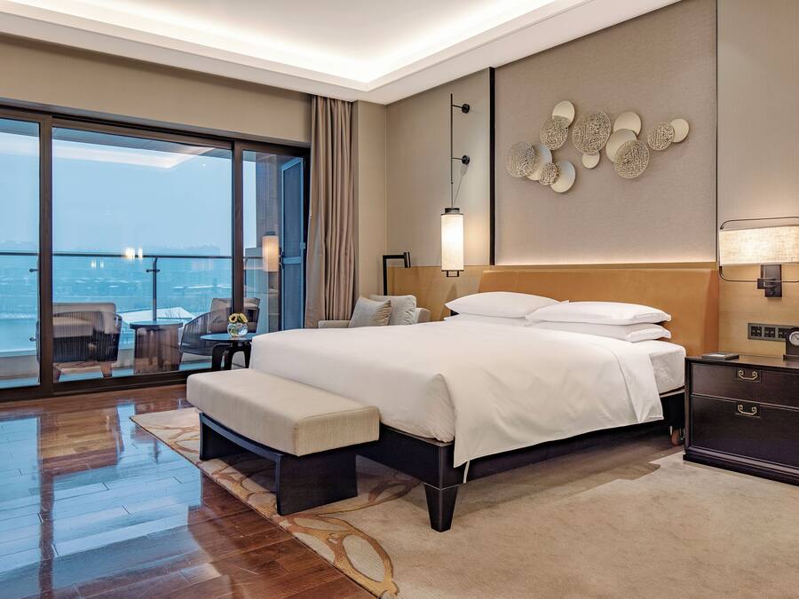 Una habitación de hotel moderna con una cama grande, una mesita de noche, un banco, vistas de la ciudad a través de ventanales del suelo al techo y un elemento decorativo en la pared.