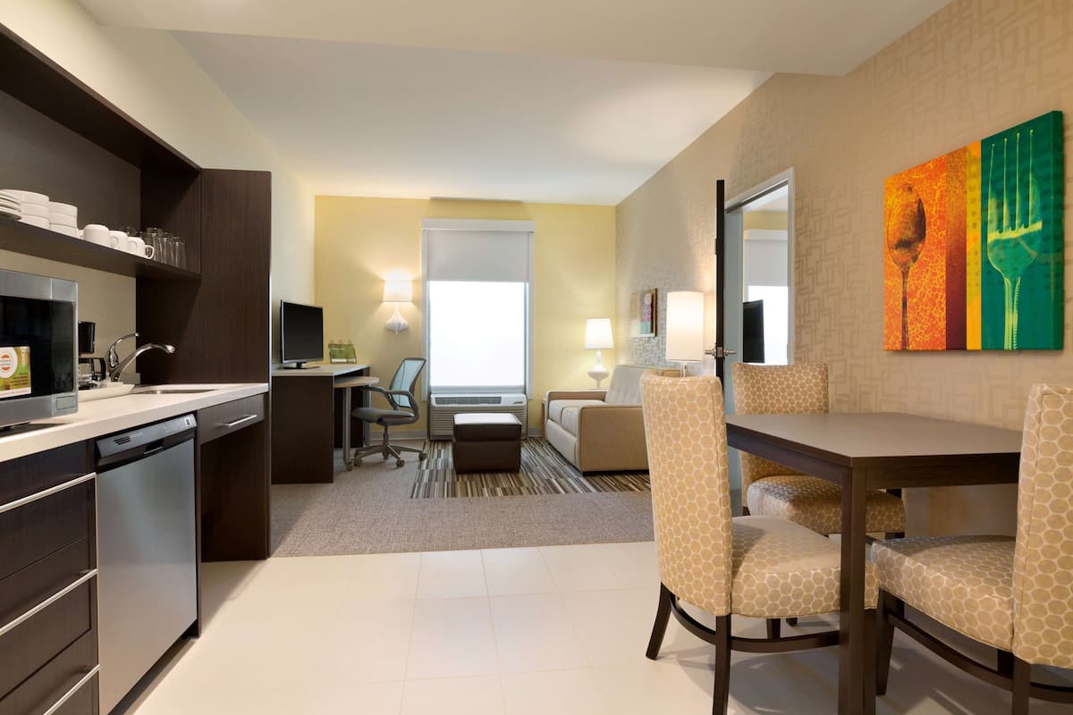 Una habitación de hotel moderna con cocina, comedor y sala de estar. Cuenta con un sofá, sillas, una mesa de comedor y obras de arte en la pared.