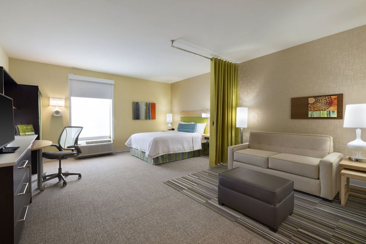 Una habitación de hotel con una cama, un escritorio con silla, un sofá, una mesita de noche con lámpara y una ventana con cortinas.