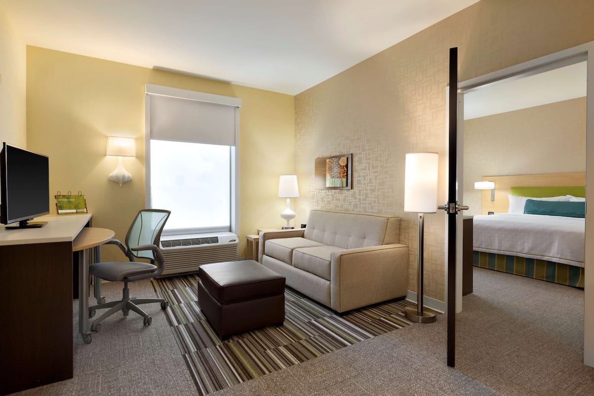 A hotel room with a desk, chair, sofa, ottoman, two bedside lamps, a bed, and a window with blinds.