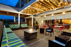 Un espace extérieur aménagé avec un foyer, des sièges confortables et une terrasse couverte dotée de poutres en bois et de guirlandes lumineuses.