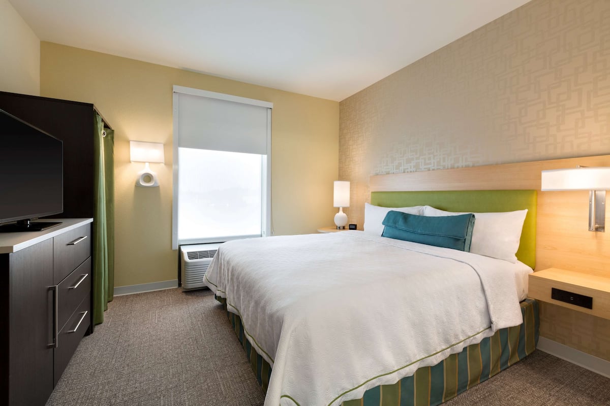Habitación de hotel con cama, televisor, ventana con persianas y lámpara de pared.