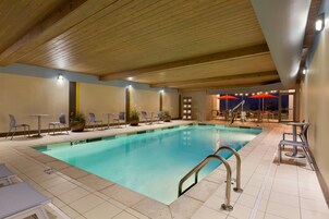 Uma piscina interior com teto de madeira, chão em azulejo e áreas de estar com mesas e cadeiras.