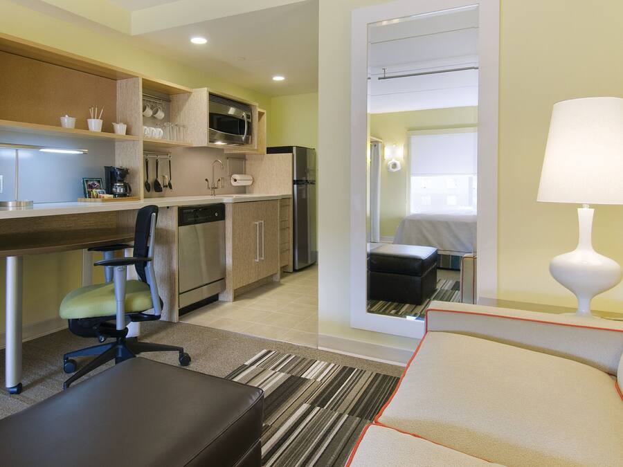 A compact hotel room with a kitchenette, a dining area, a sofa, and a lamp.