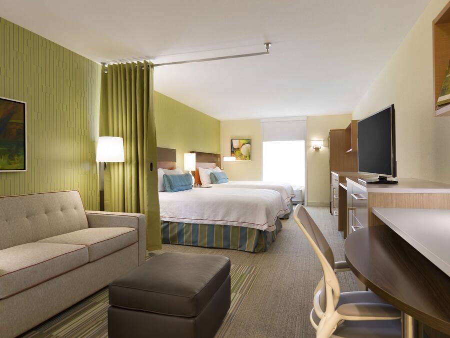 A hotel room with a sofa, a bed with two pillows, a desk with a chair, a television, and a painting on the wall.
