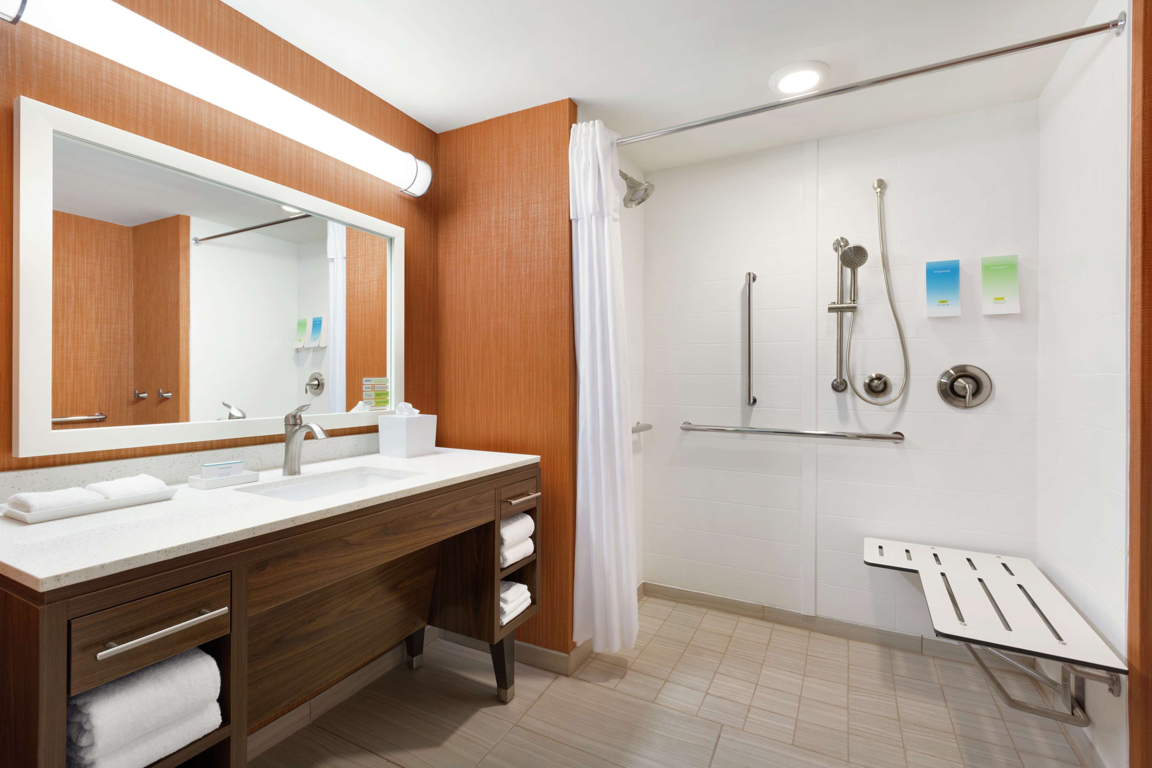 Ein modernes Badezimmer mit einem Holzbadschrank, einem großen Spiegel, einer Dusche mit Duschbrause und einer Sitzbank.