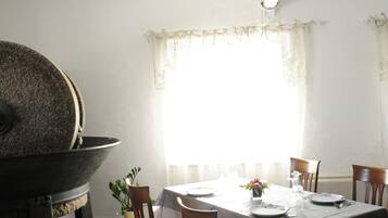 Un coin repas avec une table dressée pour quatre personnes, des chaises en bois, une grande fenêtre avec des rideaux et un plafond avec des poutres apparentes.