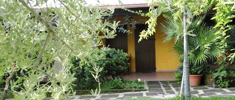 A yellow building with a dark door, surrounded by lush greenery and a stone pathway.