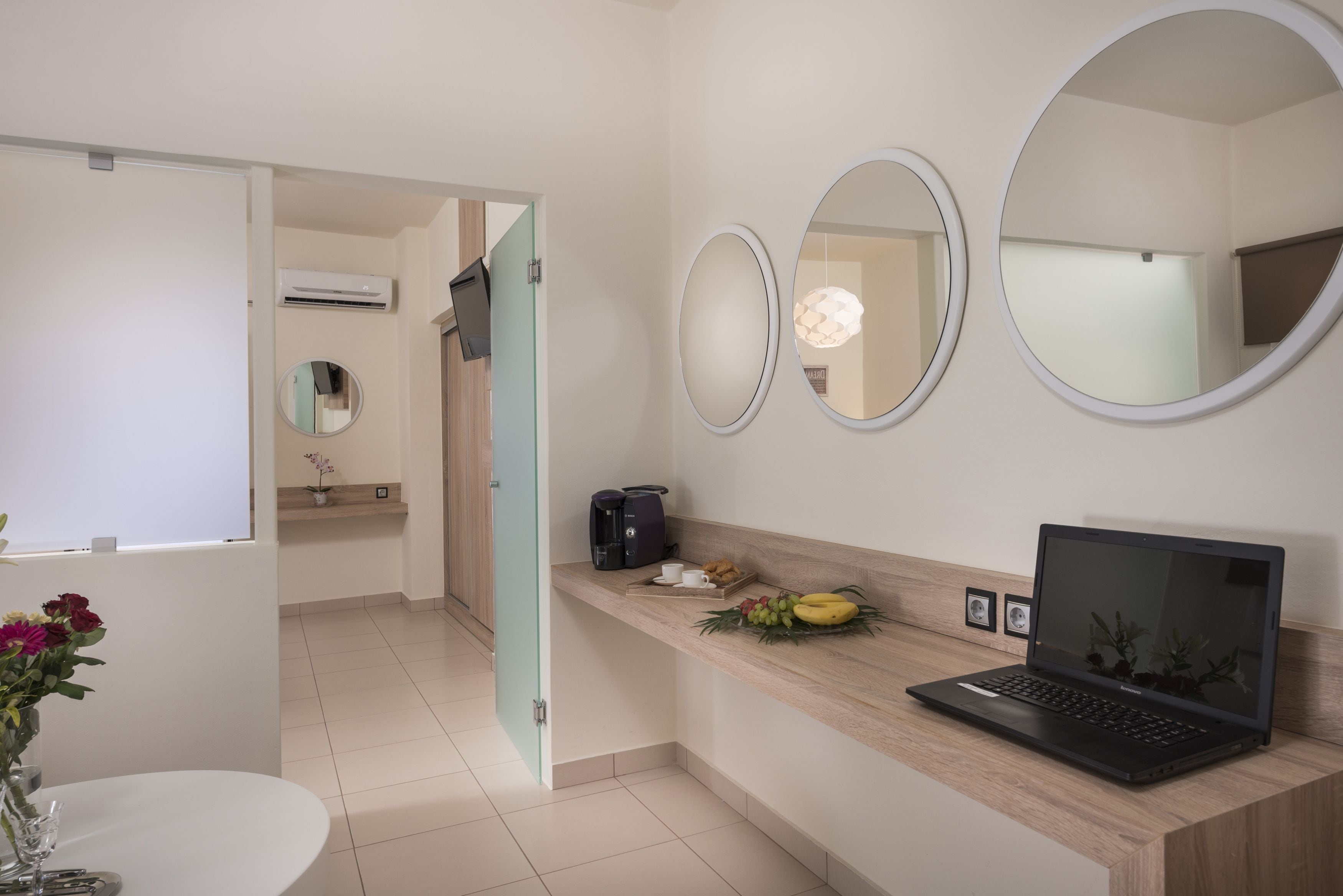 A modern bathroom with a toilet, sink, and a laptop on a vanity. There are three round mirrors on the wall, and a small table with a vase of flowers.