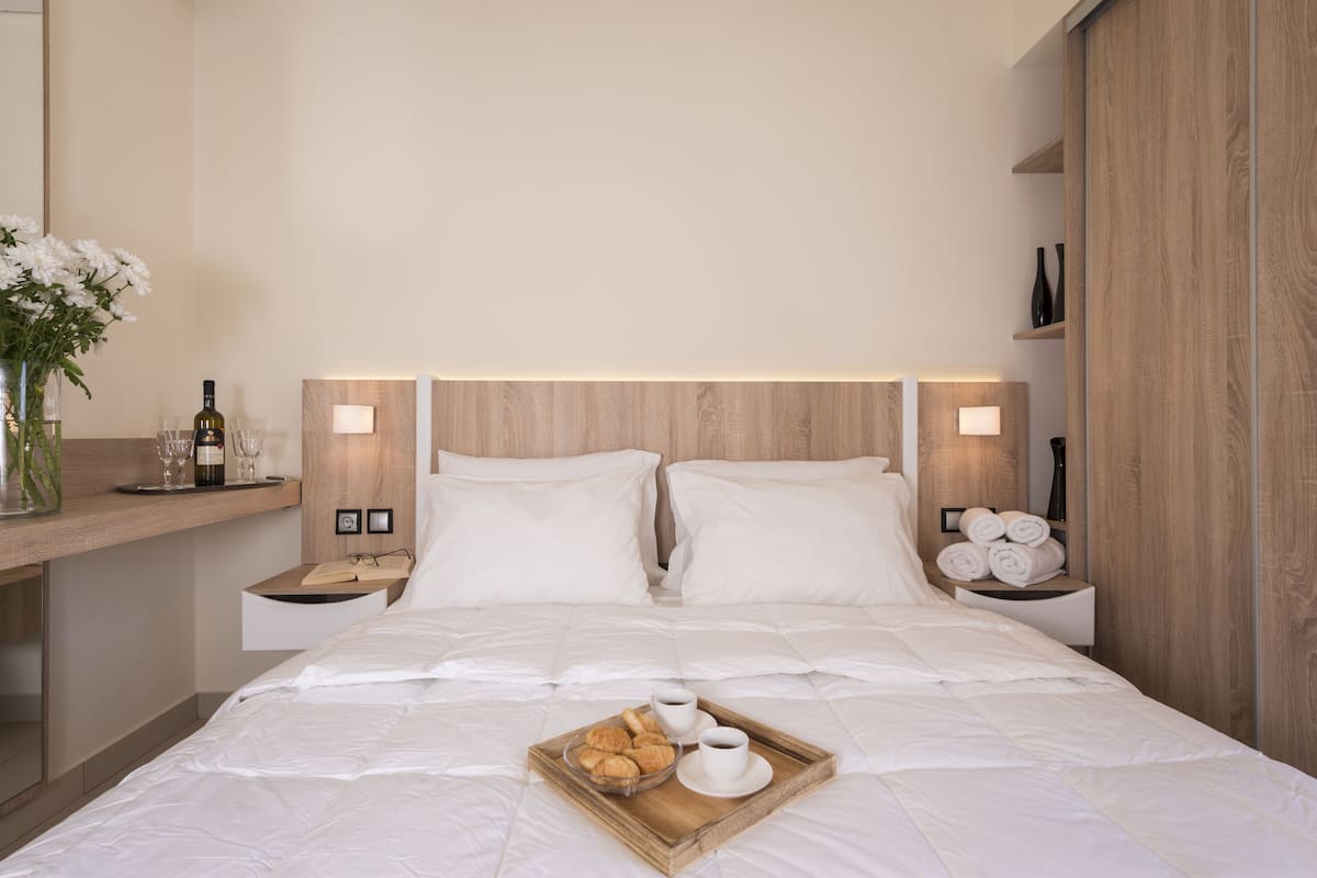 Uma cama bem arrumada com roupa de cama branca, um tabuleiro com pastéis e xícaras de café, e uma garrafa de vinho no criado-mudo.