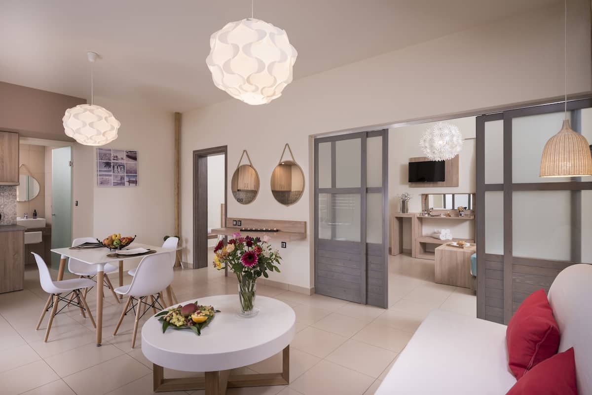 Uma sala de estar moderna com área de jantar, com uma mesa redonda, cadeiras brancas e um vaso de flores. Há duas luminárias pendentes e uma janela grande com vista.