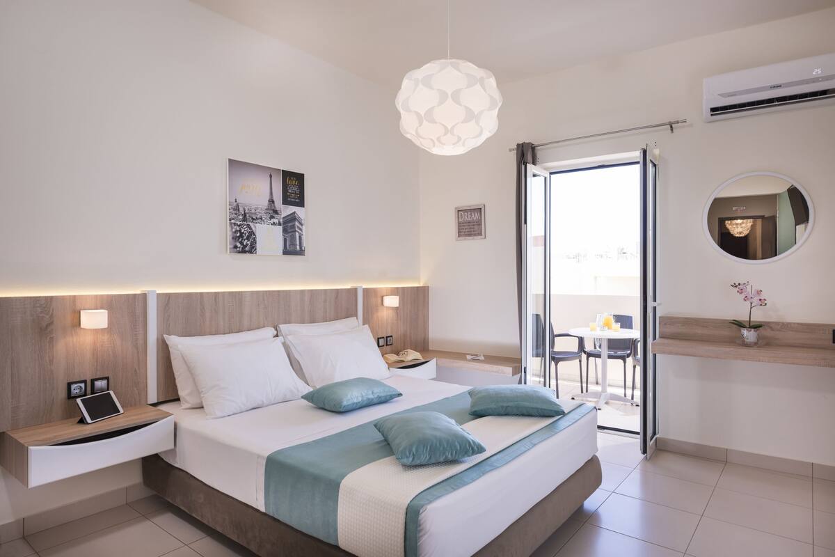 Quarto de hotel moderno com cama grande, mesas de cabeceira, luminária, ar condicionado fixado na parede, luminária de teto, quadro emoldurado e porta de vidro corrediça para uma varanda.