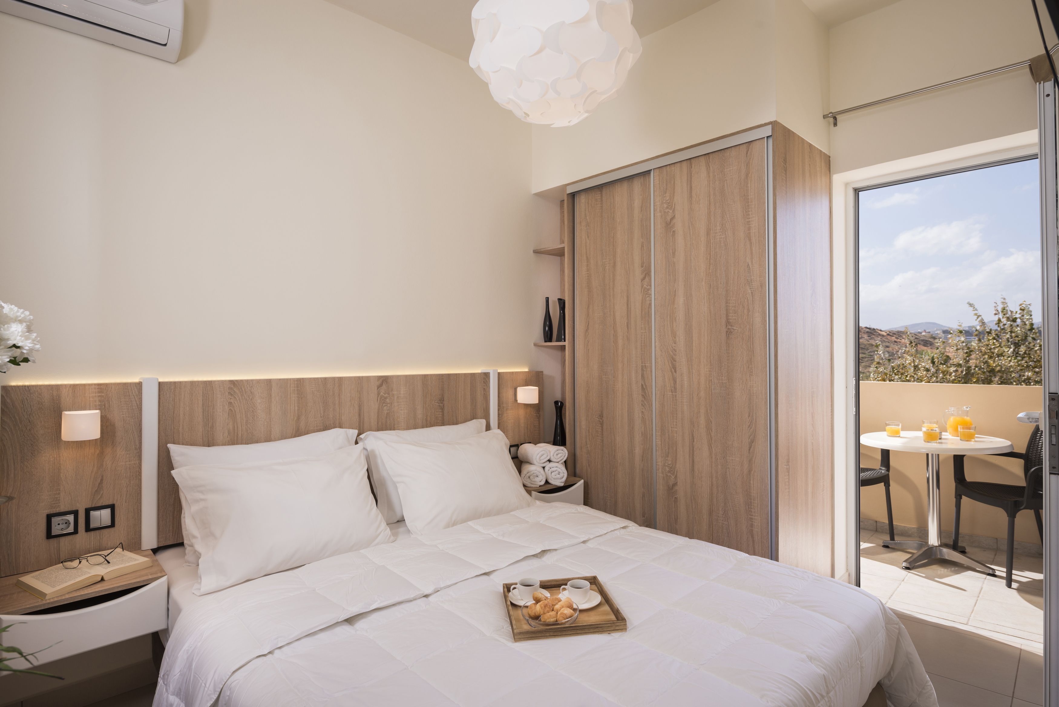 Ein modernes Hotelzimmer mit einem großen Bett, einem Nachttisch mit einer Auswahl an Gebäck, einem Beistelltisch und Blick ins Freie durch eine Schiebetür.