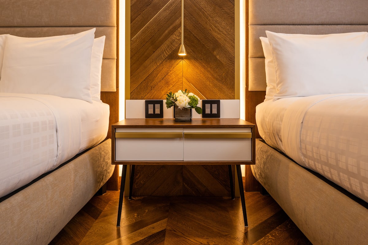 Habitación de hotel con dos camas, una mesita de noche con un florero y una pared de madera con una lámpara colgante.