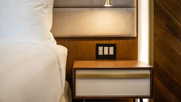 A neatly made bed with white linens, a wooden nightstand with a white drawer, and a wall socket.